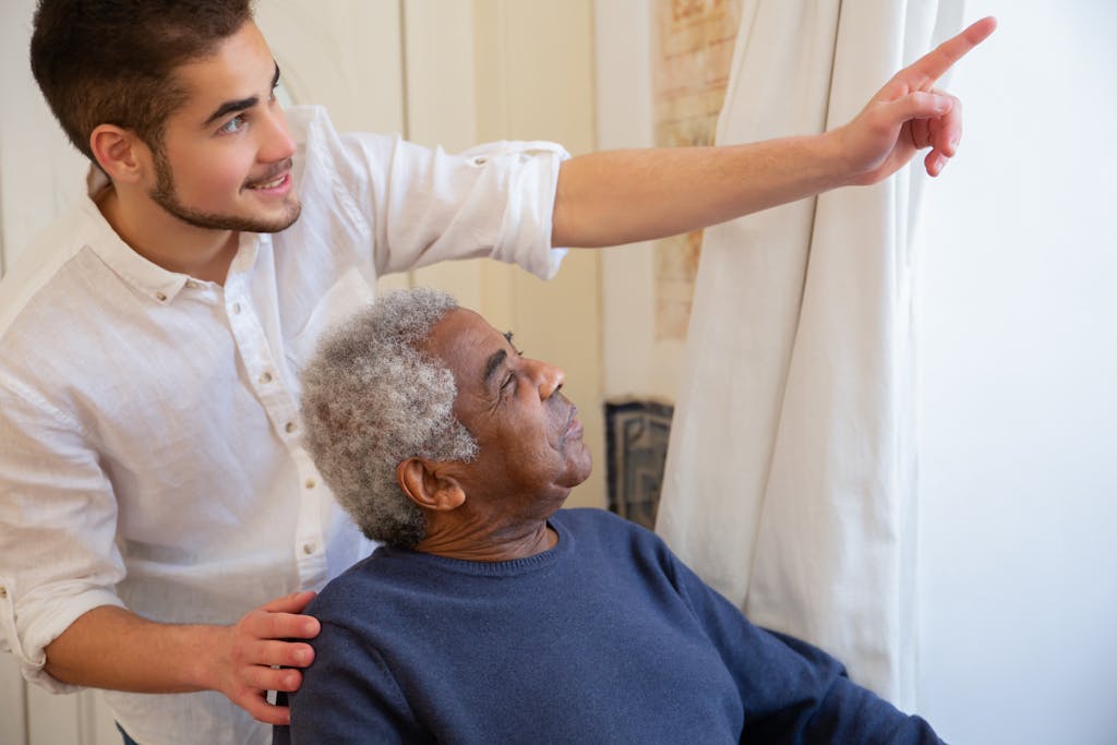 Young caregiver engaging with senior man indoors, pointing towards a window, illustrating supportive interaction in assisted living environment.